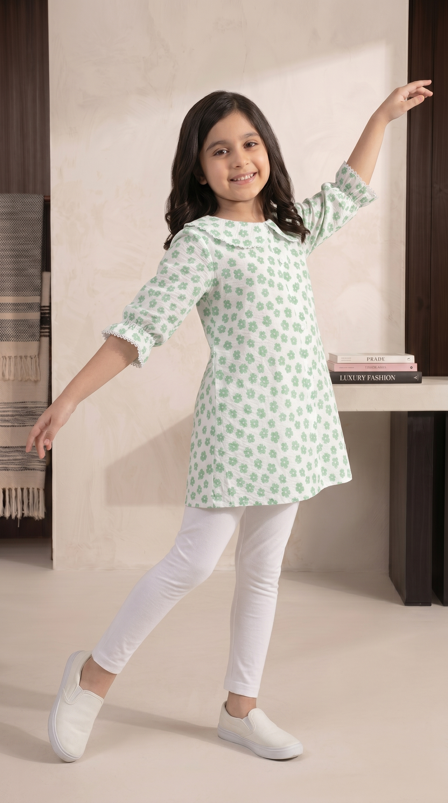Young girl wearing a green and white patterned kurta with white pants in a minimal indoor setting.